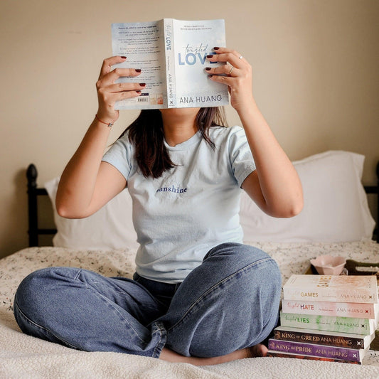 Person sitting cross-legged on bed reading Ana Huang's book Twisted Love with a stack of Ana Huang novels beside them