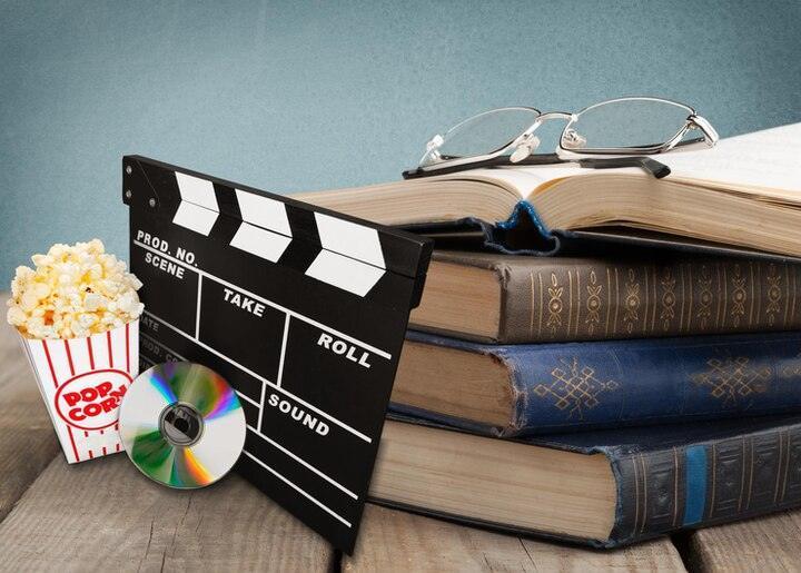 Stack of vintage books with reading glasses, film clapperboard, popcorn cup, and DVD on wooden table