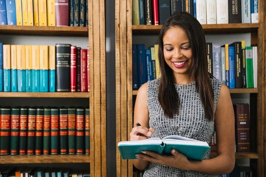 Smiling woman reading and taking notes in a library with wooden shelves filled with colorful books