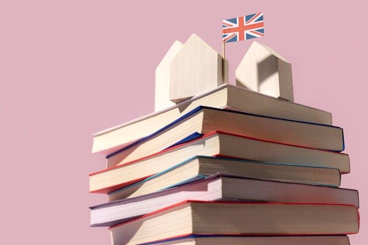 Stack of hardcover books with wooden house models and a UK flag on top against a pink background