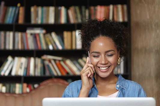 Young woman talking on phone and using laptop in cozy library with bookshelf background