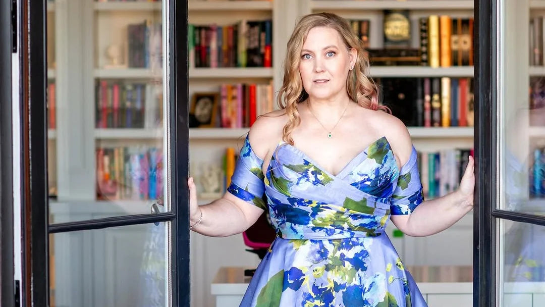 Woman in floral dress standing in front of bookshelf, fiction and self-help books visible
