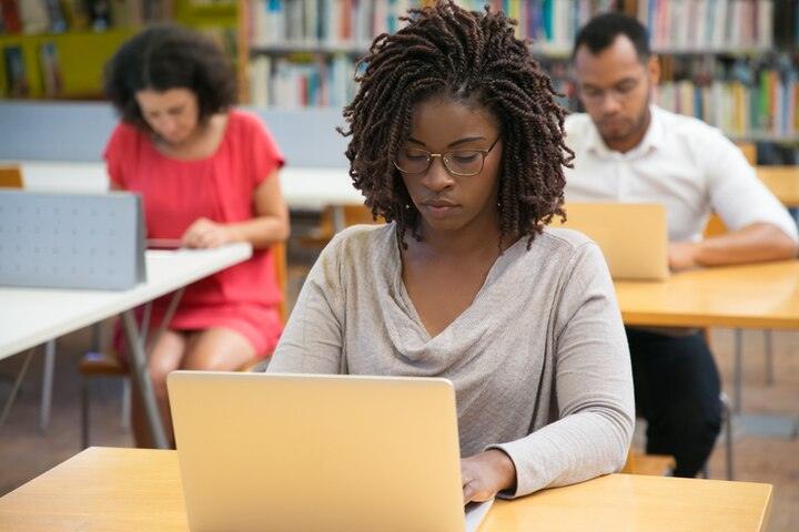 Young woman with glasses studying on a laptop in a library with two people in the background